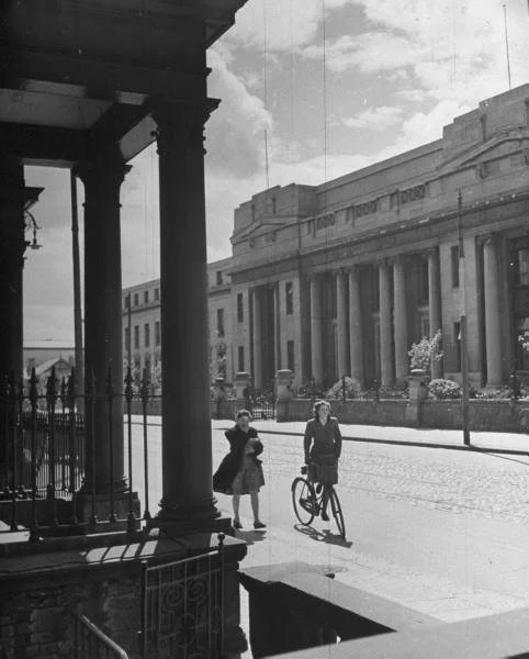 A black and white picture of people walking between 2 buildings with pillars.
